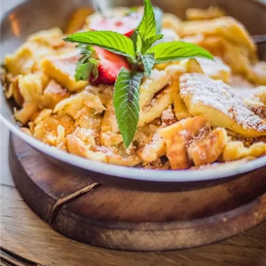 Ein Teller mit köstlichem Kaiserschmarrn, garniert mit frischen Erdbeeren und Minze. Die süße, fluffige Speise liegt auf einem Holzbrett.