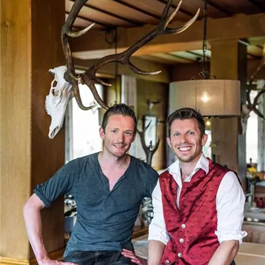 Zwei Männer posieren lächelnd in einem rustikalen Restaurant mit Holzdekor und einem Geweih im Hintergrund. Sie tragen traditionelle Trachten und präsentieren eine einladende Atmosphäre.