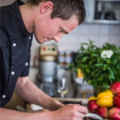 Ein Koch bereitet sorgfältig ein Gericht in einer modernen Küche vor. Im Hintergrund sind frisches Gemüse und Küchenutensilien zu sehen.