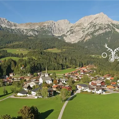 Eine malerische Landschaft mit einem kleinen Dorf und hohen Bergen im Hintergrund. Grüne Wiesen und Wälder umgeben die Gebäude.