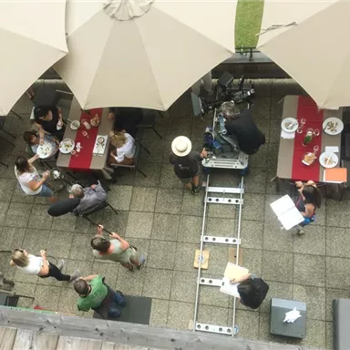 Eine Außenterrasse mit mehreren Tischen und Sonnenschirmen. Einige Gäste genießen ihr Essen, während ein Filmteam Aufnahmen macht.