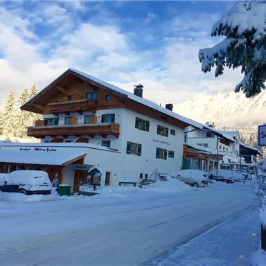 A snowy street with several houses in a winter village. The surroundings are picturesque with snow-covered trees and a clear sky.