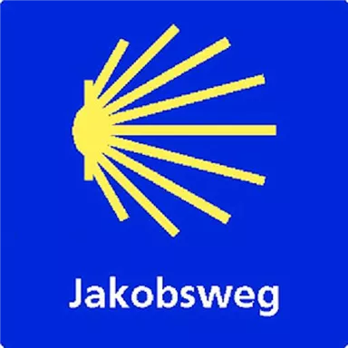 Ein blaues Schild mit einem gelben Symbol, das eine Sonne oder Strahlen darstellt. Darunter steht "Jakobsweg" in weißer Schrift.