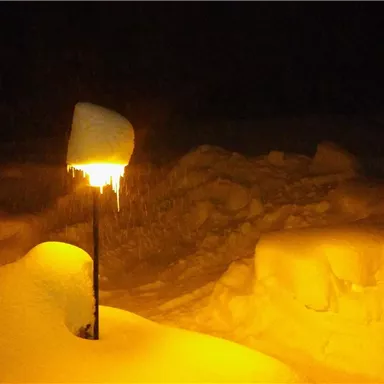A snowy landscape at night with a lit lamp. The snow is high and covers the entire ground.