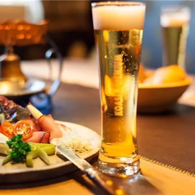 Ein Bierglas steht neben einem Teller mit Wurst, Käse, Tomaten und Gemüse. Die gemütliche Atmosphäre lädt zum Genießen ein.