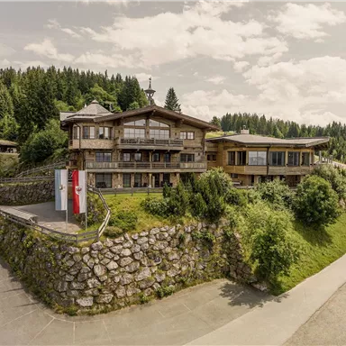 Ein modernes Holzhaus umgeben von grüner Landschaft und Bäumen. Die Straße führt sanft um das Gebäude herum.
