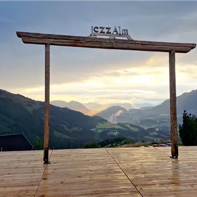 Eine aussichtreiche Plattform mit einem Holzbalken, auf dem "jazzAlm" steht. Im Hintergrund sind Berge und ein schöner Himmel bei Sonnenuntergang zu sehen.