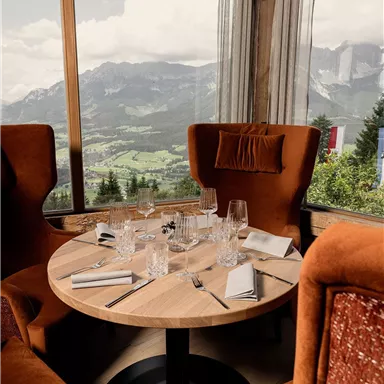 Ein stilvoller Tisch in einem Restaurant mit Blick auf die Berge. Die gemütlichen Sessel und die große Fensterfront schaffen eine einladende Atmosphäre.