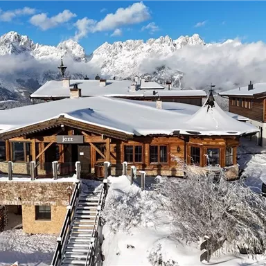 Eine malerische Berghütte in einer schneebedeckten Landschaft mit beeindruckenden Bergen im Hintergrund. Der klare blaue Himmel sorgt für eine idyllische Stimmung.