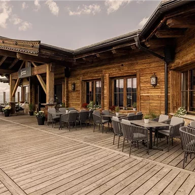 Eine gemütliche Terrasse mit Holzverkleidung und modernen Stühlen. Im Hintergrund sind große Fenster, die einen Blick auf die Umgebung bieten.