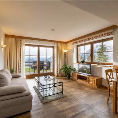 Ein gemütliches Wohnzimmer mit einem Sofa, einem Tisch und großen Fenstern. Der Raum ist hell und hat einen Blick auf die Natur draußen.