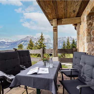 Ein gemütlicher Balkon mit einem Tisch und Stühlen. Im Hintergrund sind beeindruckende Berge und ein klarer Himmel zu sehen.