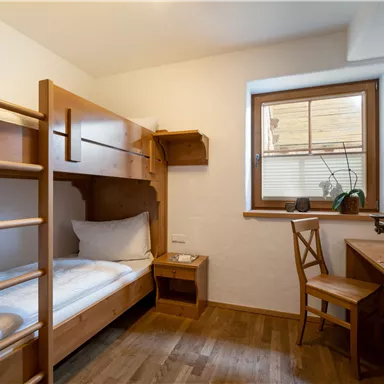 Ein einfaches Zimmer mit Etagenbetten und einem Schreibtisch. Durch das Fenster fällt sanftes Licht auf den Holzboden.