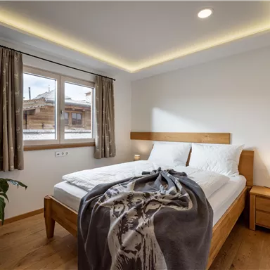 Ein gemütliches Schlafzimmer mit einem Doppelbett und einem modernen Holzdesign. Fenster lassen natürliches Licht herein und eine Pflanze sorgt für eine einladende Atmosphäre.