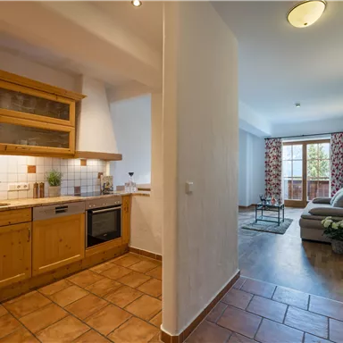 Eine moderne Küche mit Holzregalen und einer gemütlichen Sitzecke im Hintergrund. Der Raum hat fliesenboden und Zugang zu einem Balkon mit Fenster.