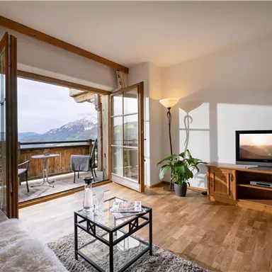 Ein gemütliches Wohnzimmer mit Holzboden und großen Fenstern. Der Blick geht auf die Berge und die Terrasse.