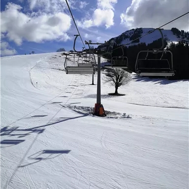 A ski lift facility on a snowy slope under a clear blue sky. In the background, some mountains and clouds are visible.