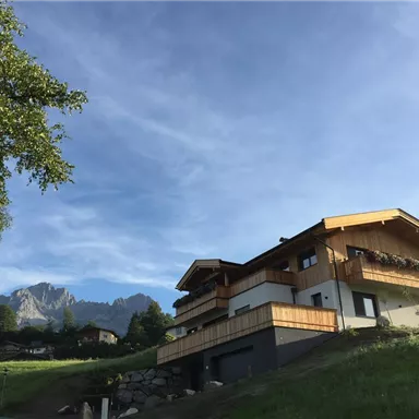 A modern house in the mountains with wooden cladding and a balcony. The landscape is green and the mountains are visible in the background.