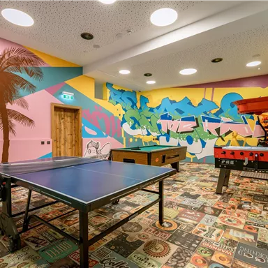 Ein Spielzimmer mit einem Tischtennistisch und einem Tischfußballspiel. Die Wände sind mit bunten Grafiken und Palmenmotiven dekoriert.