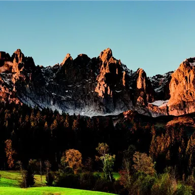 An impressive mountain landscape with rocks and a clear sky. In the foreground, green meadows and trees are visible.