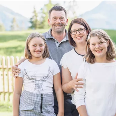 A family is standing outdoors in front of a green meadow. They are smiling kindly at the camera.