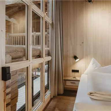 A modern bedroom with wood paneling and large windows. The decor is simple and stylish with two pillows and warm lighting.