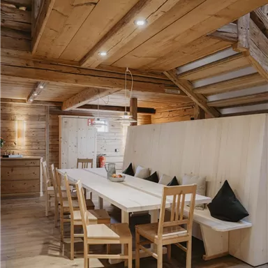 A cozy dining area with a long table and wooden seats. The interior features warm wood tones and conveys an inviting atmosphere.