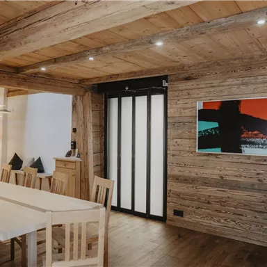 A stylishly furnished living space with wooden walls and a large dining table. A modern picture hangs on the wall, and the lighting creates a cozy atmosphere.