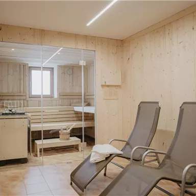 A modern relaxation room with wood paneling and two loungers. In the background is a sauna with a glass front and window.