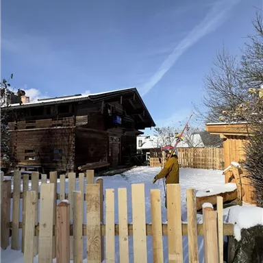 A snow-covered garden with a wooden house in the background. In the foreground stands a wooden picket fence.