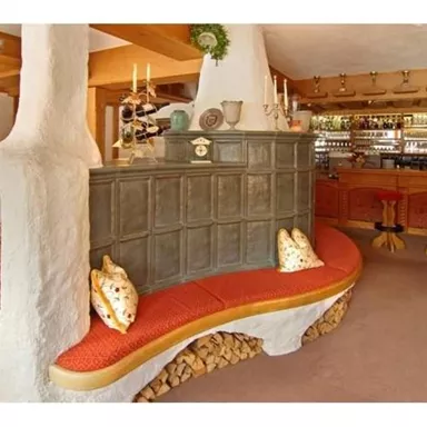 A cozy reception area with a red-upholstered bench and a wood stove. In the background, there is a bar with chairs and a warm, inviting atmosphere.
