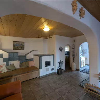 Ein gemütliches Wohnzimmer mit Holzdecke und Fliesenboden. Es gibt eine Sitzecke mit Kissen und einen Kachelofen.