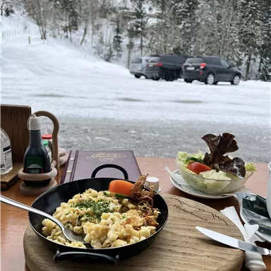 Ein gemütlicher Essensplatz mit einer heißen Pfanne voller frischer Nudeln und einem Salat. Im Hintergrund ist eine verschneite Landschaft mit Bäumen und Autos zu sehen.