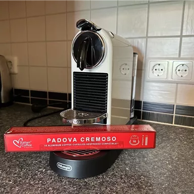 A modern coffee machine stands on a countertop, next to it lies a pack of coffee capsules with the label "PADOVA CREMOSO". The kitchen looks bright and modern.