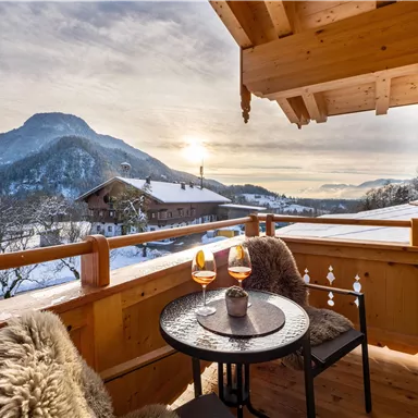 Eine gemütliche Terrasse mit Blick auf die schneebedeckten Berge. Auf dem Tisch stehen zwei Gläser, während die Sonne untergeht.
