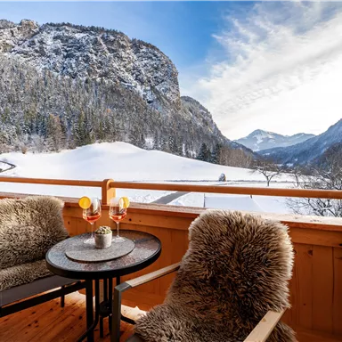 Ein gemütlicher Balkon mit zwei Stühlen und einem Tisch, umgeben von schneebedeckten Bergen. Der Himmel ist klar und die Landschaft ist malerisch.