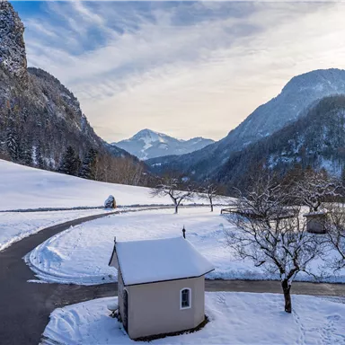 Eine verschneite Landschaft mit malerischen Bergen im Hintergrund. Ein kleiner, weißer Bau steht an einer geschwungenen Straße, umgeben von Bäumen.