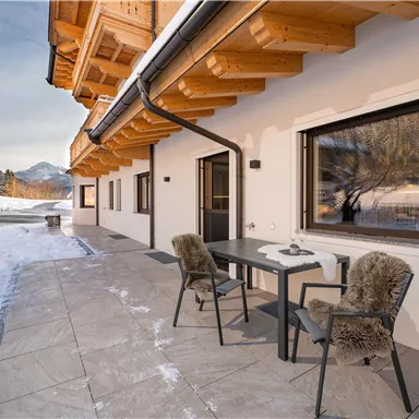 Eine moderne Terrasse mit zwei Stühlen und einem Tisch, umgeben von schneebedeckten Bergen. Die Architektur kombiniert Holz- und Steinmaterialien und schafft eine gemütliche Atmosphäre.