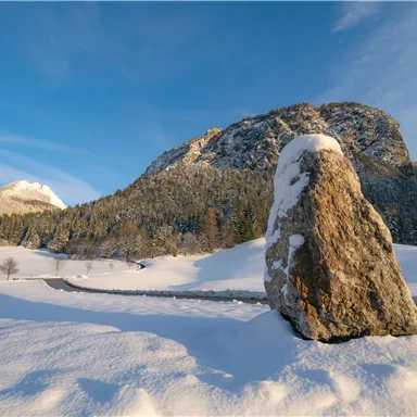 Eine winterliche Landschaft mit schneebedeckten Bergen und einem großen Stein im Vordergrund. Der Himmel ist blau und klar.