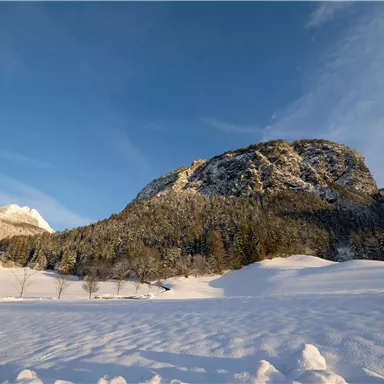 Eine winterliche Landschaft mit schneebedeckten Hügeln und einem großen Felsen im Hintergrund. Der Himmel ist klar und blau.