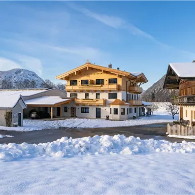 Ein charmantes alpenländisches Haus in einer verschneiten Landschaft. Im Hintergrund sind Berge und ein klarer blauer Himmel zu sehen.