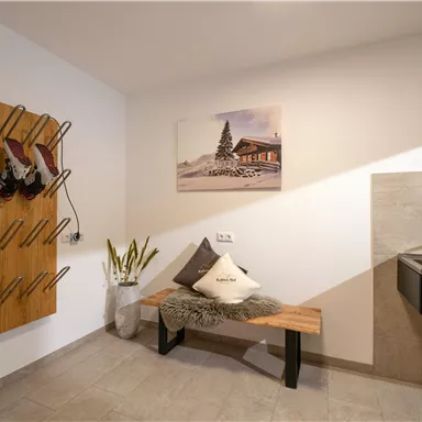 Ein moderner Eingangsbereich mit einem Schuhregal an der Wand und einem Holzbank. Im Hintergrund ist ein Waschbecken und ein Bild von einem Chalet.