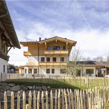 Eine moderne Almhütte mit Holzverkleidung und einem Balkon. Der Garten ist von einem niedrigen Holzzaun umgeben, umgeben von sanften Hügeln.