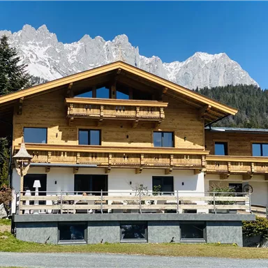A cozy wooden house with several balconies and a large terrace. In the background, impressive mountains and a clear blue sky can be seen.