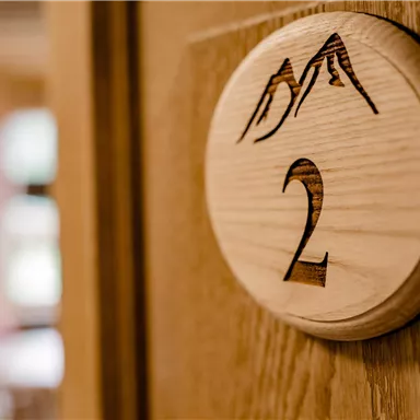 A wooden door sign with the number 2 and a mountain design. In the background, a blurred room is visible.