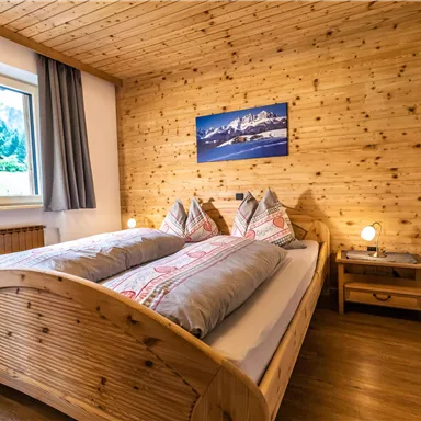 A cozy bedroom with a wooden bed and two pillows. The walls are made of wood, and a window offers a view outside.
