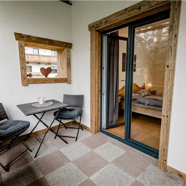 A cozy balcony with two chairs and a small table. Behind a glass door, a bedroom is visible.