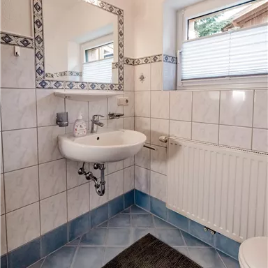 A small bathroom with white tiles and a blue floor. There is a sink, toilets, and a window with light.