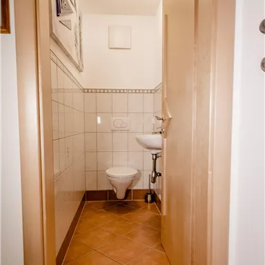 A small, clean bathroom area with a toilet and a sink. The walls are tiled and the floor is laid with brown tiles.