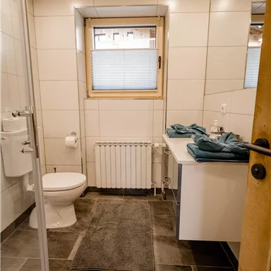 A modern bathroom with a shower, a toilet, and a sink. Towels in turquoise are ready, and a window provides daylight.